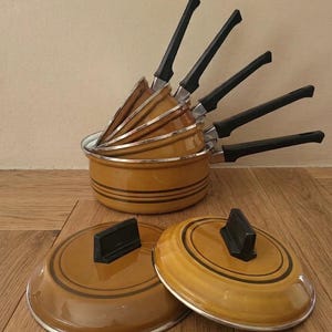 Op de afbeelding: Een set in elkaar gestapelde mosterdgele steelpannen met zwarte kunststof handgrepen en zilverkleurige randen. De pannen zijn voorzien van donkerbruine horizontale strepen aan de buitenzijde. Twee bijpassende ronde deksels liggen voor de pannen.