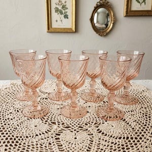 Peut inclure: Ensemble de sept verres à vin vintage teintés de rose. Chaque verre présente un motif en spirale sur le bol et un pied détaillé. Les verres sont disposés sur une nappe en crochet crème, créant une esthétique vintage.