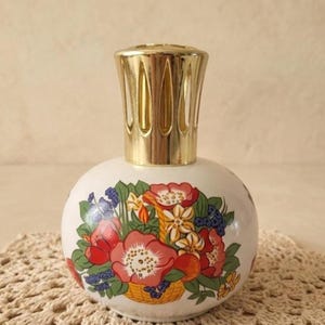 Puede incluir: Una botella decorativa blanca y redonda con una tapa dorada. La botella está adornada con un diseño floral colorido con flores rojas, rosas y azules, junto con hojas verdes. La botella es probablemente de cerámica o vidrio.