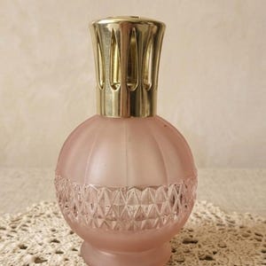 Könnte beinhalten: Eine rosa Duftlampe aus Glas mit einem goldfarbenen Metallaufsatz. Die Lampe hat einen runden Körper mit einer strukturierten, rautenförmigen Basis und vertikalen Rippen. Der Aufsatz ist ein dekoratives, geschlitztes Design.