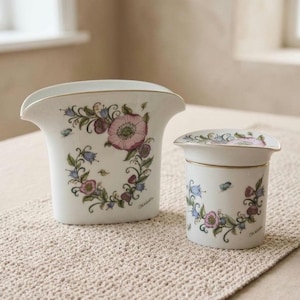 Peut inclure: Deux vases en céramique blanche avec des motifs floraux. Le plus grand vase a un dessus évasé et une fleur rose avec des feuilles vertes. Le plus petit vase est cylindrique avec un couvercle. Les deux ont une bordure dorée.