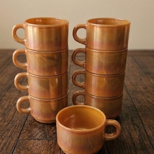Op de afbeelding: Set van acht amberkleurige koffiekopjes met oren. De kopjes zijn gestapeld in twee kolommen van vier, met één kopje apart geplaatst. De kopjes hebben een glanzende afwerking en staan op een donker houten oppervlak.