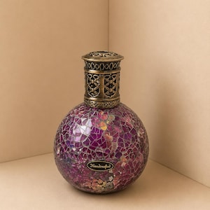 Könnte beinhalten: Eine kugelförmige Duftlampe mit einem violetten Mosaikglaskörper aus irisierenden Scherben. Die Oberseite besteht aus einer dekorativen, perforierten, messingfarbenen Metallkappe mit Filigranmuster. Ein kleines ovales Etikett zeigt den Text Handcrafted.