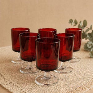 Peut inclure: Six verres à vin rouge rubis avec un bol court et cylindrique et un pied et une base transparents. Les verres sont disposés sur une surface texturée de couleur beige. Les verres sont en verre rouge foncé.