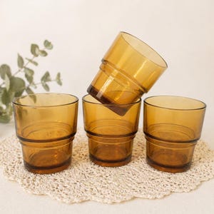 May include: Four amber-colored drinking glasses. The glasses have a cylindrical shape with a ridged design around the middle and base. One glass is tilted, resting on the others. The glasses are on a cream-colored, crocheted doily.