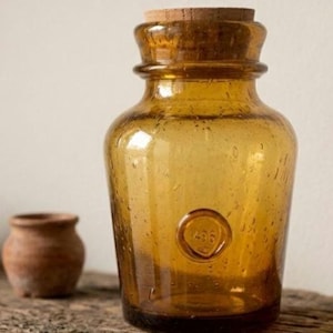 May include: An amber-colored glass jar with a wide neck and a cork stopper. The glass features small air bubbles throughout and a circular embossed detail on the front containing the number 1436. The jar has a rounded body and a thick rim.