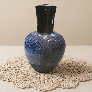 May include: A decorative blue and white speckled ceramic fragrance lamp with a black top. The lamp sits on a beige crocheted doily. The lamp has a rounded body and a tall, cylindrical top.