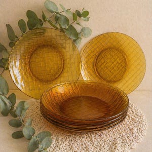 Peut inclure: Ensemble d'assiettes en verre ambré avec une texture tissée. Les assiettes sont disposées sur un dessous de plat en crochet crème, avec des feuilles d'eucalyptus en arrière-plan. Idéal pour une table chaleureuse.