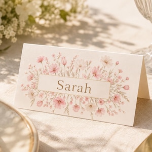 May include: A folded white place card featuring a central rectangular box with the name Sarah printed in gold lettering, surrounded by a delicate watercolor illustration of pink and white wildflowers and green foliage on the front panel.