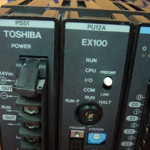 May include: Close-up of a black Toshiba EX100 power module. The module has various labels and indicators, including "POWER", "RUN", and "24Vdc". It features multiple ports and a series of numbered lights. The text "TOSHIBA" is prominently displayed.