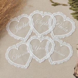 Peut inclure: Six cartons en forme de cœur avec des bordures en dentelle blanche et des noms personnalisés en écriture cursive. Les noms Jackie, Sophie, Paola, Dianne, Yasmin et Iridania sont visibles. Les cartes sont disposées sur une surface de ton neutre.