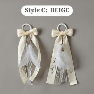 May include: Two beige ribbon ornaments labeled Style C: BEIGE, each featuring a white ring, a lace bow, white feathers, and a bronze chain charm. One ribbon displays the text Mrs Taylor and the date 08.24.27 in black script lettering.