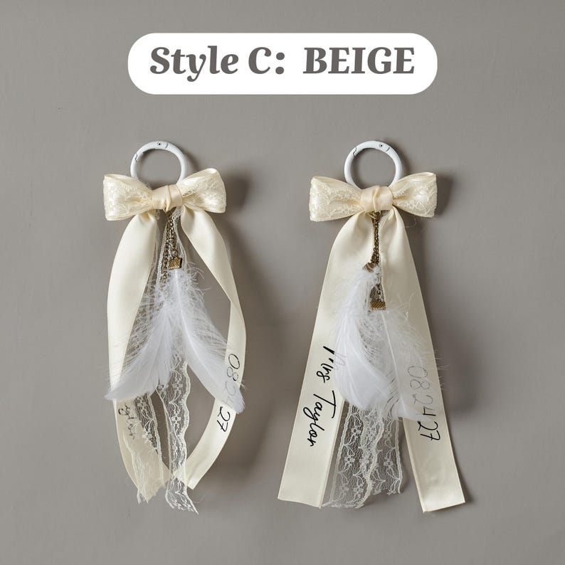 May include: Two beige ribbon ornaments labeled Style C: BEIGE, each featuring a white ring, a lace bow, white feathers, and a bronze chain charm. One ribbon displays the text Mrs Taylor and the date 08.24.27 in black script lettering.