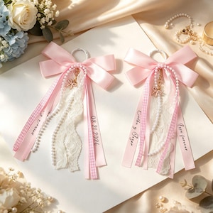 May include: Two decorative pink satin ribbon bows featuring white lace trim, pearl bead strands, and small gold-tone bow charms. Each bow includes hanging ribbons printed with the text 08.21.2026, Erika y Tezor, and Love forever, attached to a white metal ring.