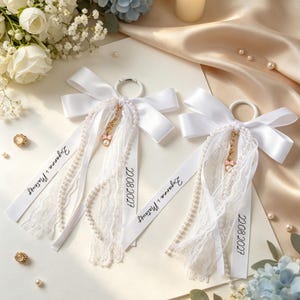 May include: Two white satin ribbon bows attached to circular rings, featuring hanging strands of white lace, pearl strings, and a small gold-tone charm with a pink bow. The ribbons are printed with the text Zuzanna i Mateusz and 22.08.2027.