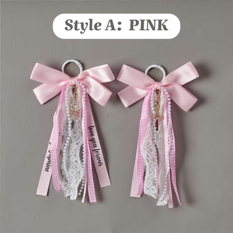 May include: Two pink satin bow keychains with white lace, gingham ribbons, and pearl strands. Each features a gold-tone bow charm and a white metal ring. One ribbon is printed with the text 'love you forever' in black script lettering.