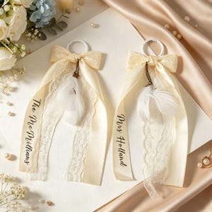 May include: Two cream-colored satin ribbon bows with white lace trim, each attached to a white circular ring. The ribbons feature white feathers hanging from a small chain and black text reading The McMillians and Mrs Holland respectively.