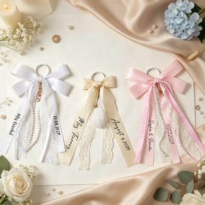 May include: Three decorative ribbon keychains featuring white, cream, and pink satin bows with lace trim, pearl strands, and metal rings. Each ribbon is personalized with black text including names like Mrs. Kiechel, Mrs. Tierney, Sasha & Randu, and dates.