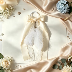 May include: A decorative accessory featuring a cream-colored satin ribbon bow attached to a white circular ring. Two soft white feathers hang from a small bronze-toned chain suspended from the center of the bow, accompanied by delicate lace trim.