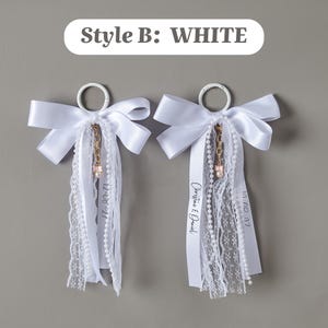 May include: Two white decorative rings featuring satin bows, lace trim, and pearl strands. A gold-toned chain with a small pink charm hangs from each ring. One ribbon displays the text "Christine & Daniel" and dates are printed on others.