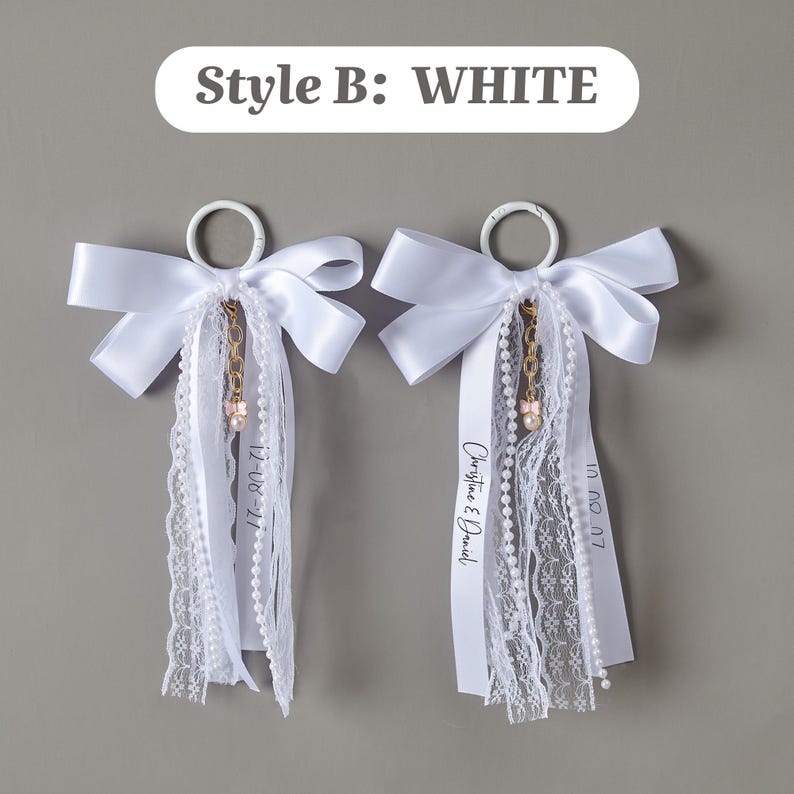 May include: Two white decorative rings featuring satin bows, lace trim, and pearl strands. A gold-toned chain with a small pink charm hangs from each ring. One ribbon displays the text "Christine & Daniel" and dates are printed on others.