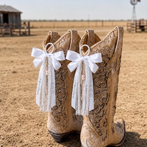 May include: A pair of tan leather cowboy boots featuring intricate embroidery and rhinestone embellishments. Each boot is adorned with a white satin ribbon bow, white lace trim, a small pearl bead, and a tag printed with Christine & Daniel 10.09.07.