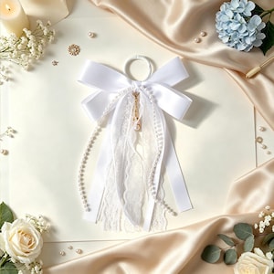 May include: A white satin ribbon bow attached to a circular ring, featuring cascading white lace strips and two long strands of small white faux pearls. A small gold-toned metal charm hangs from the center of the bow, adding a subtle metallic detail.