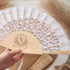 May include: A handheld folding fan featuring a white lace canopy and light wood ribs. The wood is printed with a crest containing the initials RS, the names Lucy & Sean, and the date 05.14.25. The handle is inscribed with Thank you for celebrating with us!