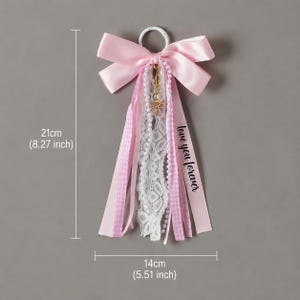 May include: A decorative hanging ornament featuring a large pink satin bow attached to a white ring. It includes cascading ribbons of pink gingham, white lace, and a string of white pearls. A small gold bow charm hangs from the center. Text reads "love you forever". Measures 8.27 inches by 5.51 inches.