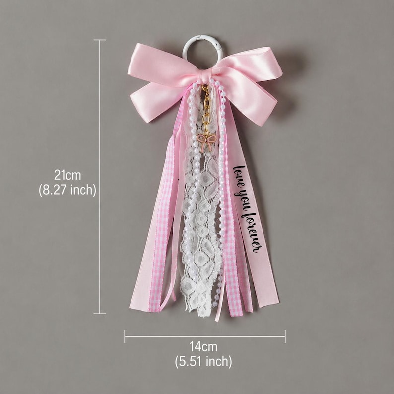 May include: A decorative hanging ornament featuring a large pink satin bow attached to a white ring. It includes cascading ribbons of pink gingham, white lace, and a string of white pearls. A small gold bow charm hangs from the center. Text reads "love you forever". Measures 8.27 inches by 5.51 inches.