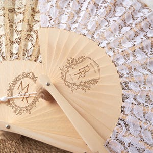 May include: A folding hand fan featuring light wood ribs and white lace fabric. The wood is engraved with two distinct designs: one with the initials M&R inside an ornate frame, and another with the initials BR, the text KENNY & MIA, and 05.14.26.
