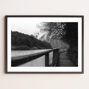 Può includere: Fotografia in bianco e nero incorniciata di un paesaggio. Una passerella in legno con ringhiera costeggia un corso d'acqua. Alberi e una collina sono visibili sullo sfondo, con un cielo nuvoloso.