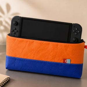 Op de afbeelding: Een rechthoekige draagtas voor een draagbare spelcomputer, voorzien van een feloranje bovengedeelte en een donkerblauw ondergedeelte. De tas heeft een klein rood lipje aan de zijkant en een klein logolabel. Een zwarte spelcomputer is in de tas geplaatst.