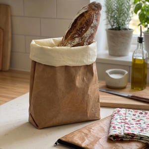 Puede incluir: Una bolsa de pan de papel marrón con un interior color crema contiene una barra de pan crujiente. Una bolsa plana a juego con cremallera negra y un paño con estampado floral están junto a la bolsa de pan. La escena está ambientada en una cocina.