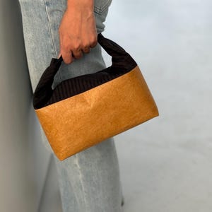 Pode incluir: Uma pequena bolsa com formato retangular. A bolsa tem um exterior castanho claro e uma seção superior e alça pretas e texturizadas. A bolsa está sendo segurada por uma pessoa usando jeans azuis.