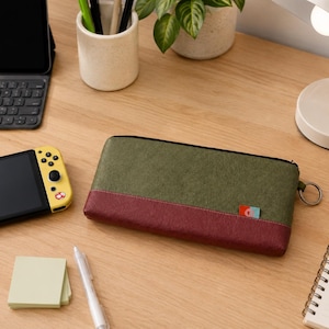 May include: A rectangular zippered pouch featuring a two-tone design with an olive green upper section and a dark maroon lower section. The pouch includes a small metal ring on the side and a tiny rectangular tag with a blue and red graphic.