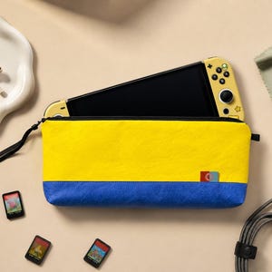 May include: A rectangular zippered pouch featuring a bright yellow upper section and a solid blue lower section. A yellow handheld gaming console is partially inserted into the pouch. Three small game cartridges, a charging cable, and earbuds are visible.