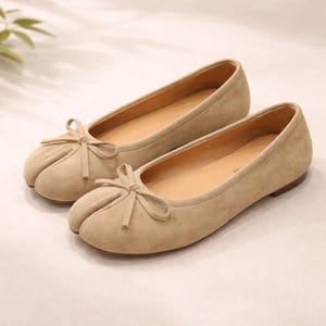 Könnte beinhalten: Ein Paar beige Ballerinas mit kleinen Schleifen auf den Zehen. Die Schuhe haben ein abgerundetes Zehendesign und eine flache Sohle. Die Innenseite des Schuhs ist hellbraun. Die Schuhe sind aus einem weichen, Wildleder-ähnlichen Material.