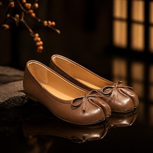 May include: A pair of brown leather ballet flats with small bows on the toes. The shoes have a rounded toe design and a smooth, polished finish. The interior of the shoes is a lighter tan color.