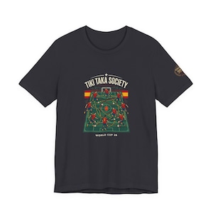 May include: Dark gray t-shirt featuring a soccer field graphic with players in red and yellow uniforms. The text "TIKI TAKA SOCIETY" is above the field, and "WORLD CUP 26" is below. A small emblem is on the sleeve.
