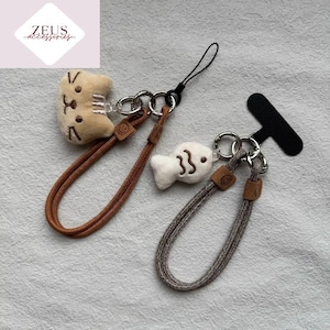 Puede incluir: Dos colgantes para teléfono con diseños de animales de peluche. Uno presenta una cara de gato beige, el otro un pez blanco. Ambos tienen cordones marrones y grises, anillos metálicos y detalles de cuero. También se ve un agarre negro para el teléfono.