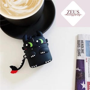 May include: Black silicone AirPod case shaped like a cartoon dragon with green eyes and white accents. The dragon has a tail with red tips. The case is on a white surface next to a black coffee cup and a newspaper.