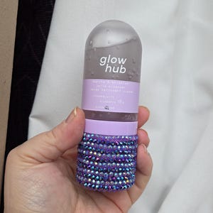 May include: A clear bottle of Glow Hub Purify & Brighten Jelly Cleanser. The bottle has a purple base covered in sparkling purple gems. The bottle has a clear top with the words "glow hub" in white.