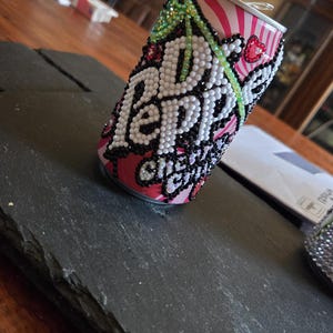 May include: A decorated Dr. Pepper Cherry soda can. The can is embellished with white, black, and green rhinestones forming the Dr. Pepper logo. The can has a pink and white striped pattern with red heart details.