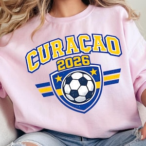 May include: A light pink sweatshirt with the word "CURACAO" in yellow and blue lettering, and the year "2026". A soccer ball graphic is centered below the text, with blue and yellow accents. The sweatshirt is worn by a person.