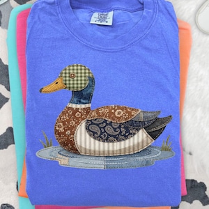 May include: A stack of t-shirts in blue, pink, and orange. The top shirt is blue and features a patchwork duck design with various patterned fabrics. The duck has a green and brown checkered head, brown body, and blue and white striped wings.