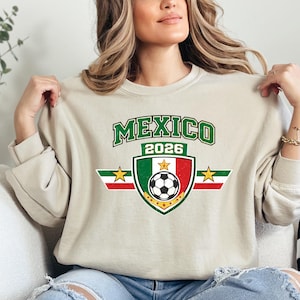 May include: Beige sweatshirt with a green and white "MEXICO 2026" graphic. The graphic includes a shield with the Mexican flag colors, a soccer ball, and stars. The sweatshirt has long sleeves and a crew neck.