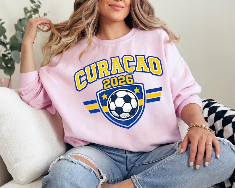 May include: A light pink sweatshirt with the word "CURACAO" in yellow and blue lettering, and the year "2026". A soccer ball graphic is centered below the text, with blue and yellow accents. The sweatshirt is worn by a person.