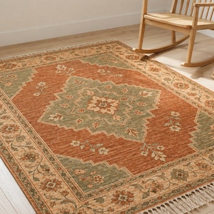May include: A rectangular area rug featuring a central sage green diamond medallion on a terracotta orange field, surrounded by a floral border with cream accents and finished with short, light-colored fringe along the two shorter ends.