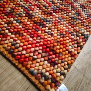 Op de afbeelding: Een kleurrijk tapijt met een textuur van kleine, ronde, viltwollen ballen. Het tapijt heeft een mix van rode, oranje, bruine en blauwe tinten, met een goudbruine rand. Het tapijt ligt op een houten vloer.
